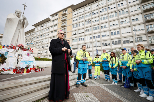 En biskop ber rosenkransen sammen med en gruppe kvinner og menn ved Johannes Paul II-statuen utenfor Gemelli-sykehuset