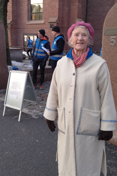 En eldre kvinne ikledd ytterkåpe står i gaten utenfor St. Olav domkirke og smiler mot kamera