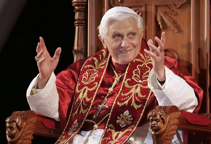 Pave Benedikt XVI i røde klær, sitter i en stol, smiler og løfter hendene