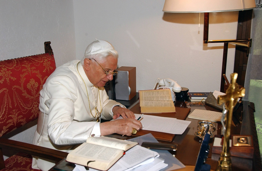 Pave Benedikt XVI, kledd i hvitt, sitter ved et skrivebord og jobber med en tekst